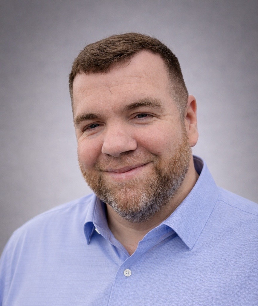 Smiling man with short brown hair and beard wearing a light blue button-down shirt, against a gray backdrop.