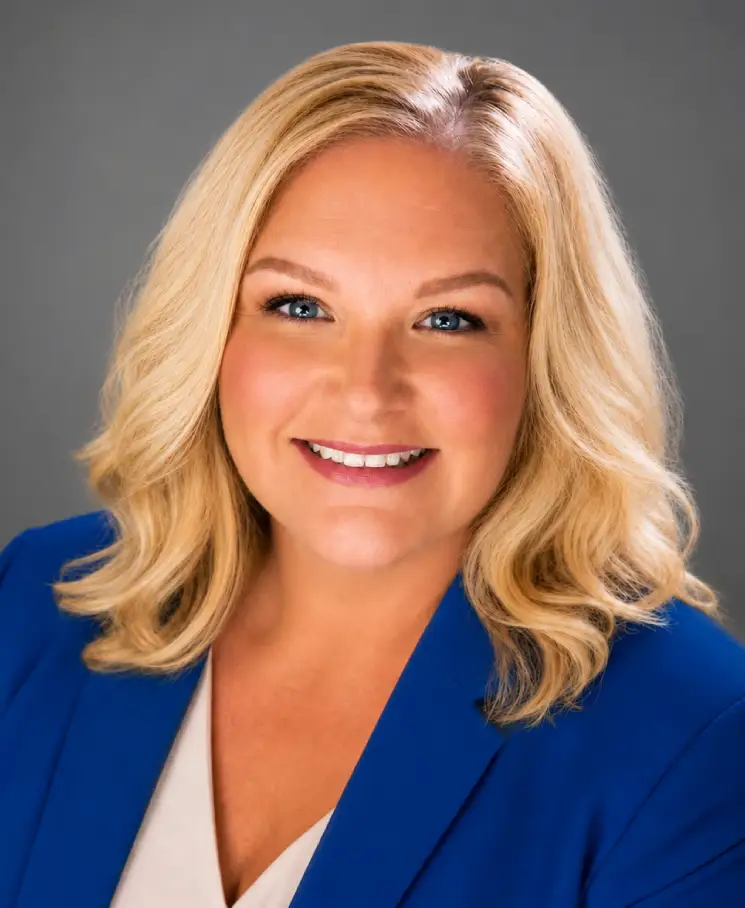 Professional headshot of a smiling blonde woman wearing a blue blazer against a gray backdrop.