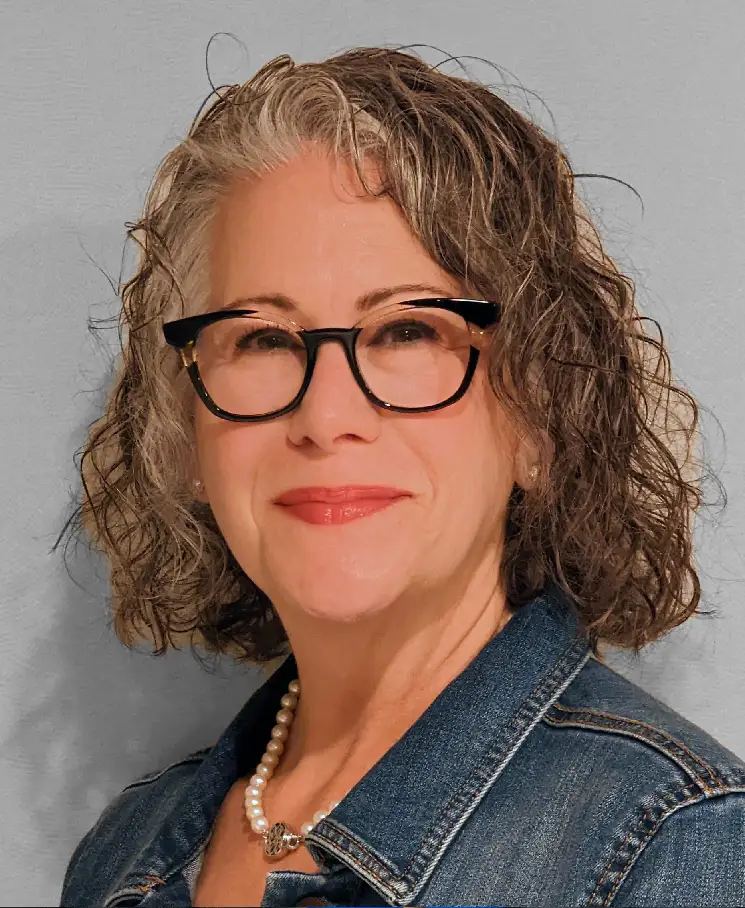 Portrait of a smiling middle-aged woman with short curly gray-brown hair, black glasses, a denim jacket, and a pearl necklace.