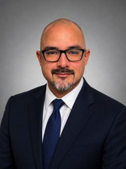 Professional headshot of a middle-aged man with glasses, wearing a navy suit and blue tie against a gray backdrop.