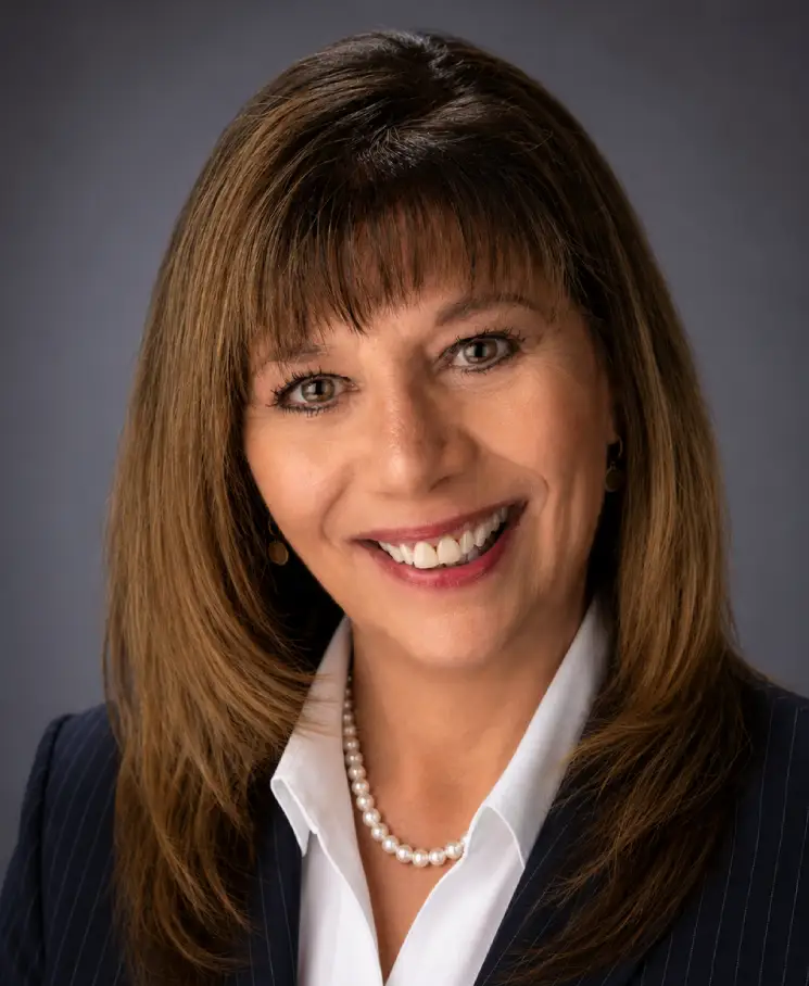 Professional headshot of a smiling woman in a navy blazer and white blouse, wearing a pearl necklace.
